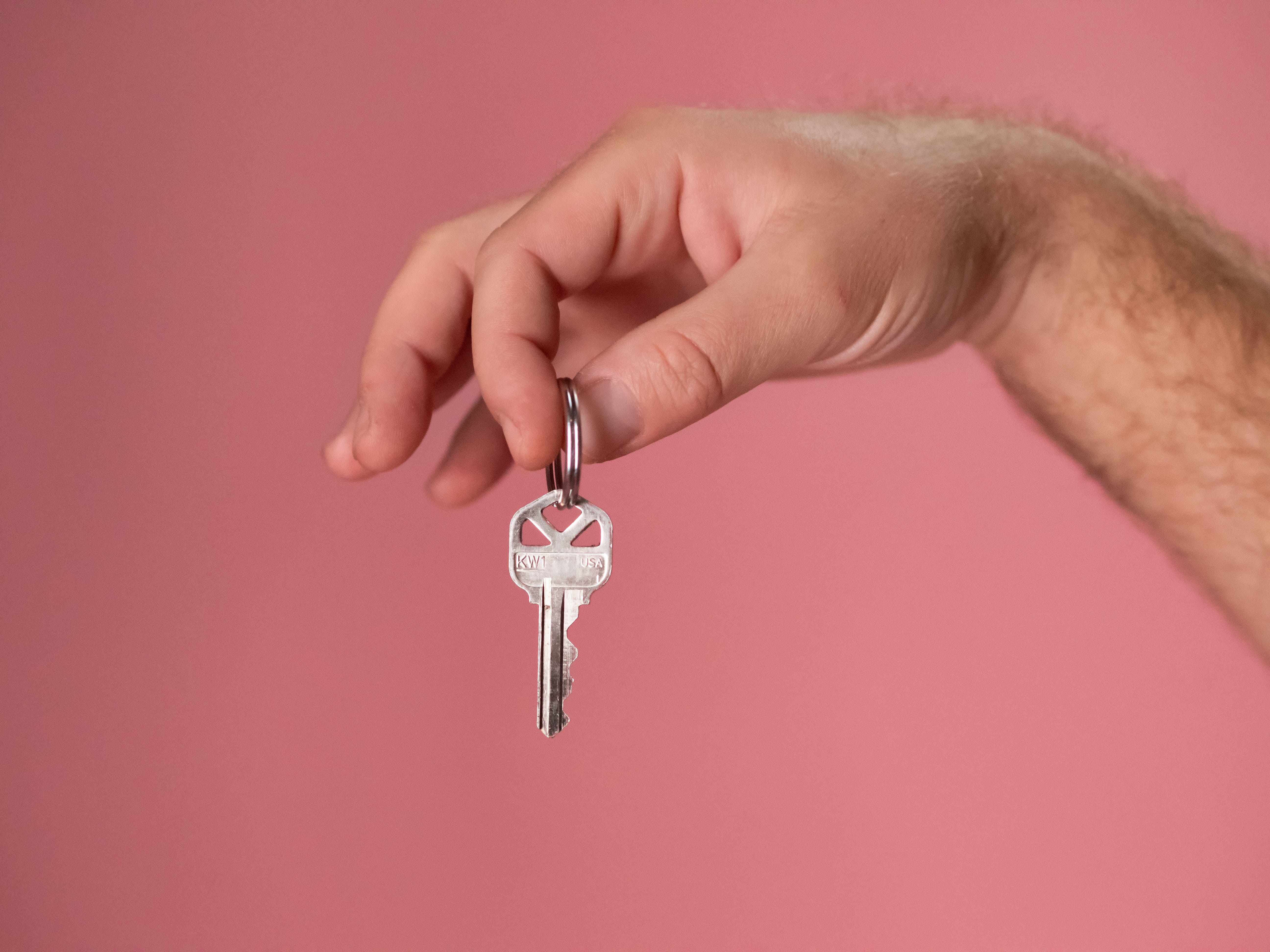 A close-up image of a person's right hand, with light skin tone, holding a silver key with a bow labeled 'KW1'. The key is displayed prominently against a plain pink background, filling the central area of the frame. This visual metaphorically represents the concept of security and access, akin to the process of learning how to create and use SSH keys for secure remote access.